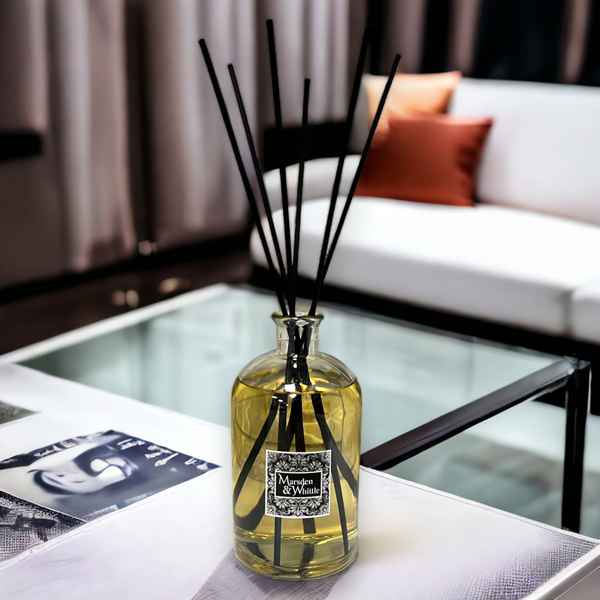 A large glass reed diffuser bottle sitting on a glass table with a cosy lounge in the background. 6 extra long black reeds are in the diffuser and the bottle is full.