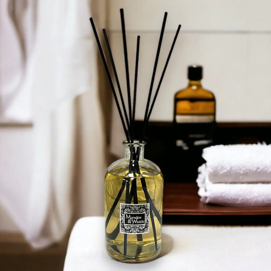 Reed diffuser with black sticks in a glass bottle on a surface, with a blurred background of towels and a bottle.