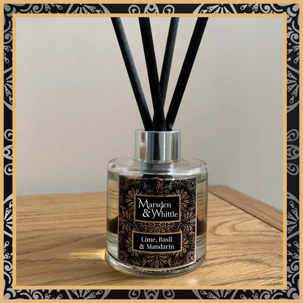 A Lime, Basil and Mandarin glass reed diffuser with black reeds and a silver chrome cap sitting on a wooden table.