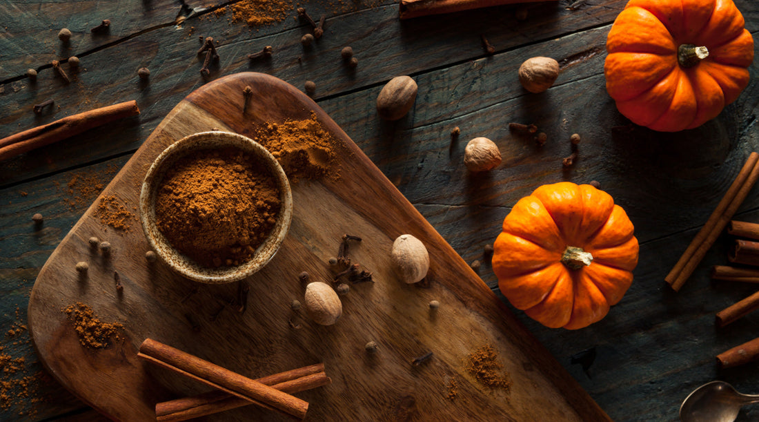 Overhead view of autumn spices on a dark rustic wooden table, with a bowl of ground pumpkin spice, cinnamon sticks, whole nutmeg and cloves on a wooden board beside two small orange pumpkins.