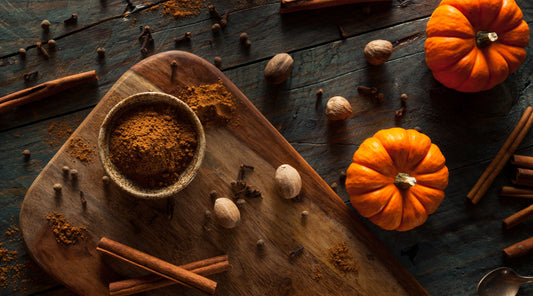 Overhead view of autumn spices on a dark rustic wooden table, with a bowl of ground pumpkin spice, cinnamon sticks, whole nutmeg and cloves on a wooden board beside two small orange pumpkins.
