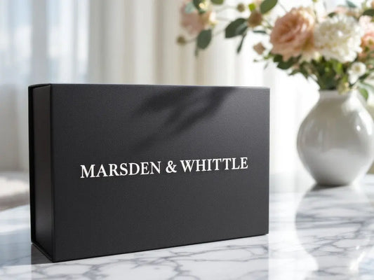 A black gift box with the wording "Marsden & Whittle" foiled onto the lid, sat on a white marble countertop with a vase of white and peach coloured peony flowers in the background,