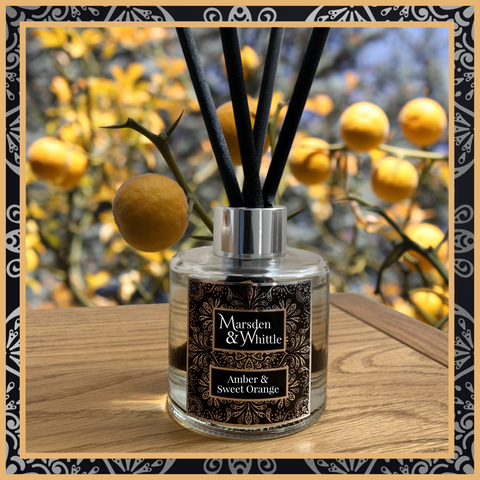 An Amber and Sweet Orange glass reed diffuser with black reeds and a silver chrome cap sitting on a wooden table.