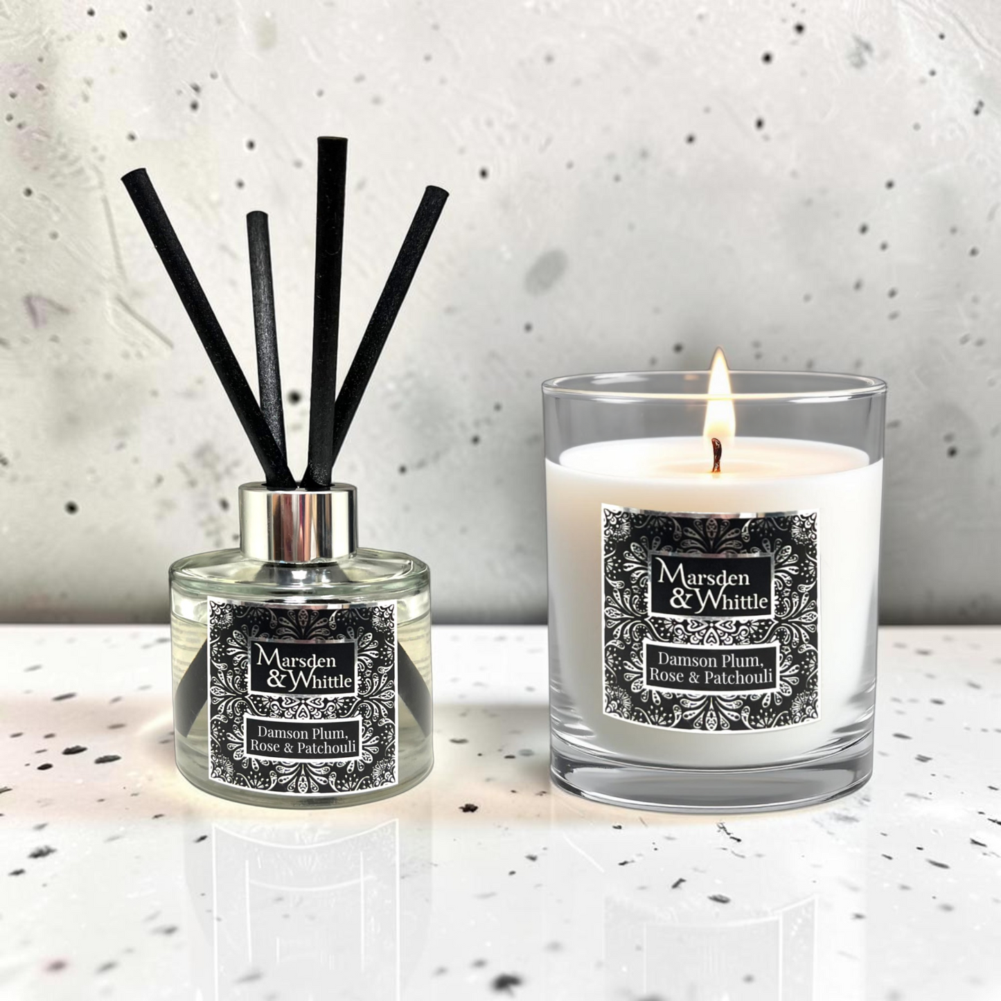 A reed diffuser and a lit scented candle from the brand Marsden & Whittle, both labeled 'Damson Plum, Rose & Patchouli,' displayed on a light speckled surface against a textured light gray wall.