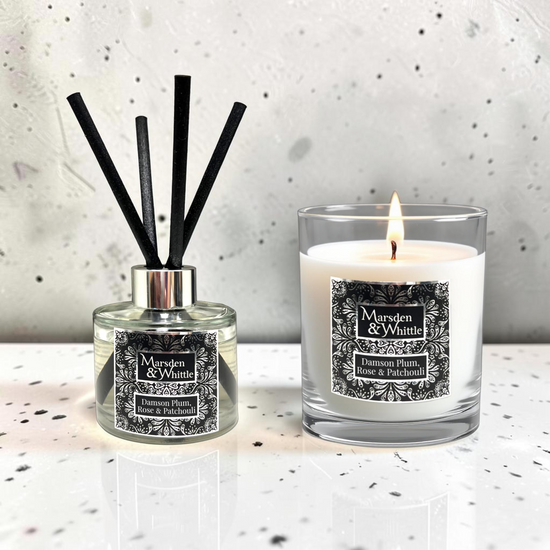 A reed diffuser and a lit scented candle from the brand Marsden & Whittle, both labeled 'Damson Plum, Rose & Patchouli,' displayed on a light speckled surface against a textured light gray wall.