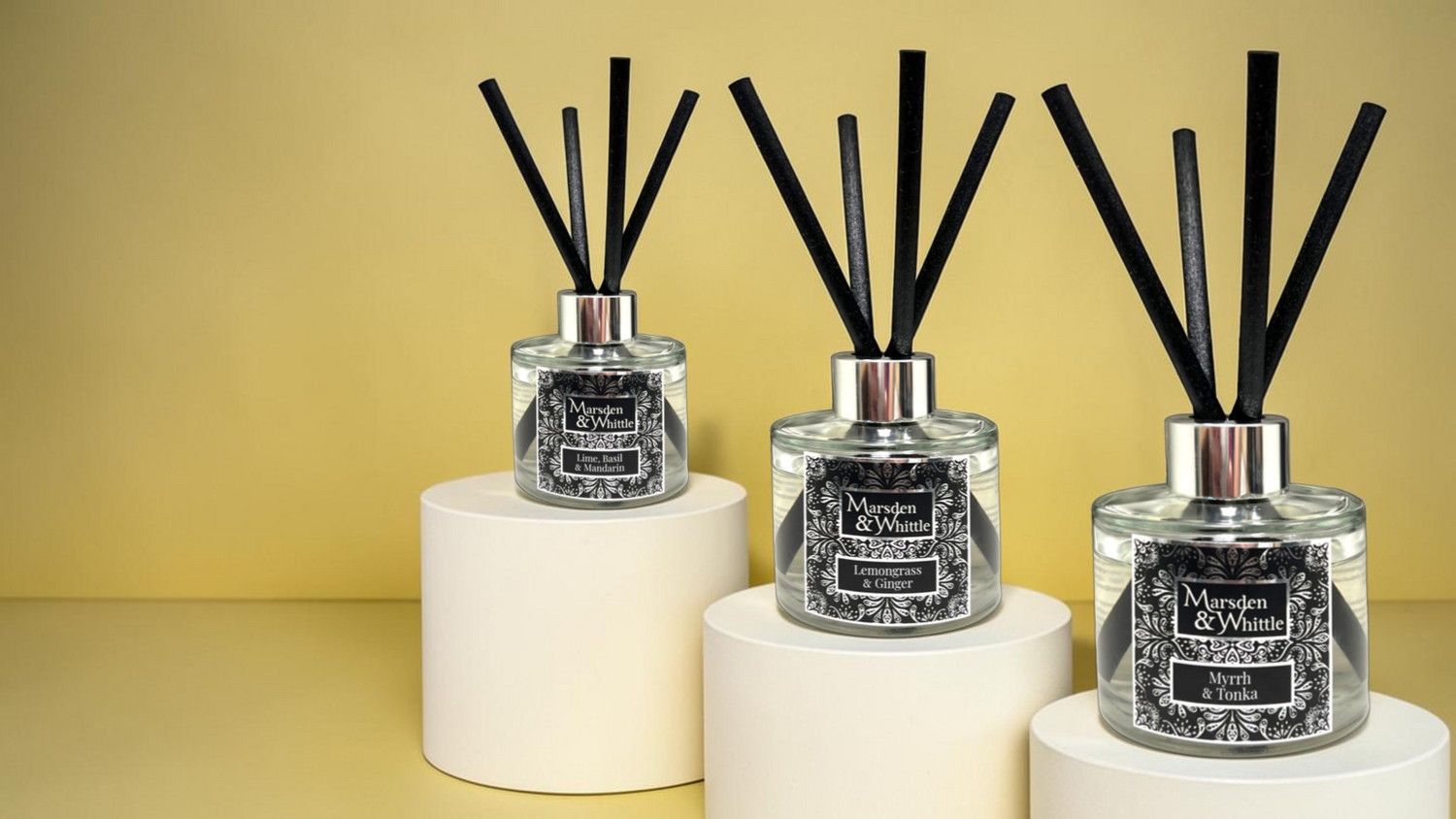 Three Marsden & Whittle Reed Diffusers setting on white plinths, each at a staggered height, against a light yellow-gold background.