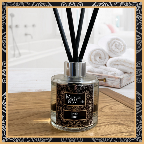 A Fresh Linen glass reed diffuser with black reeds and a silver chrome cap sitting on a wooden table.