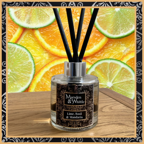 A Lime, Basil and Mandarin glass reed diffuser with black reeds and a silver chrome cap sitting on a wooden table.