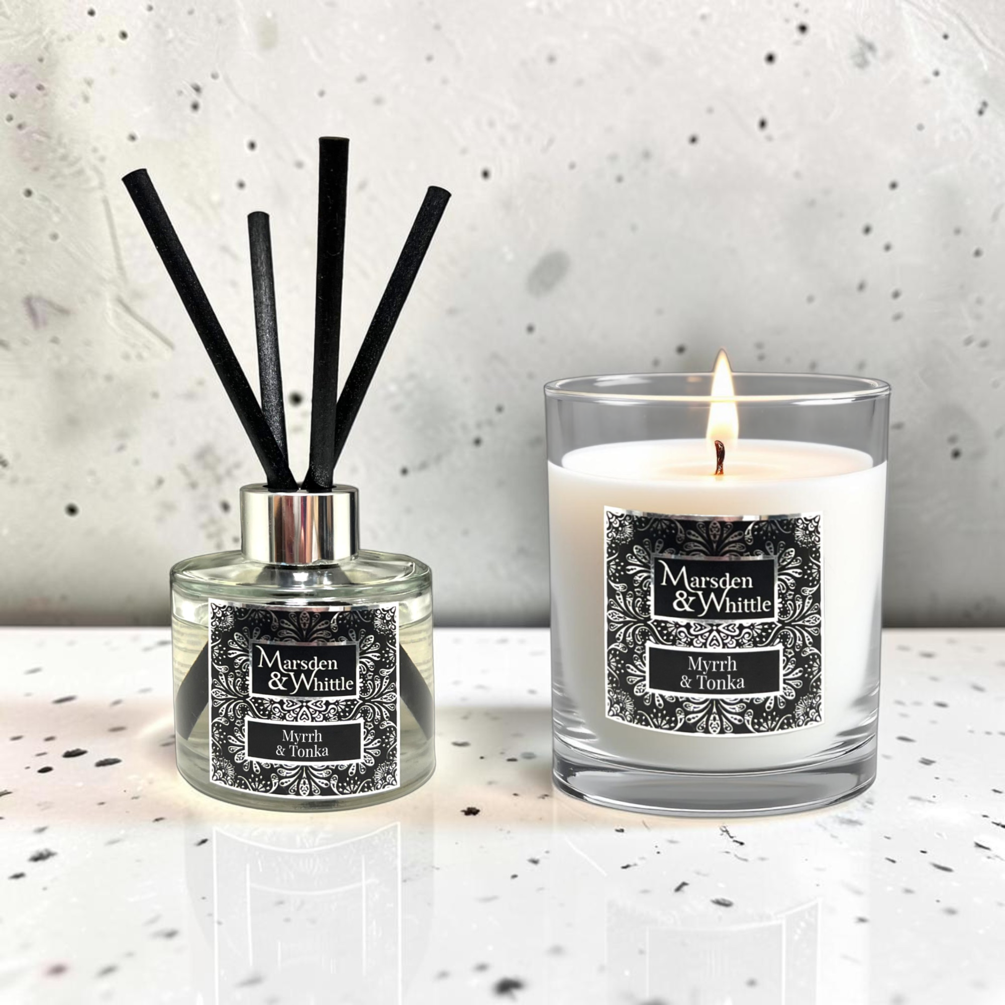 A reed diffuser and a lit scented candle from the brand Marsden & Whittle, both labeled 'Myrrh & Tonka' displayed on a light speckled surface against a textured light gray wall.