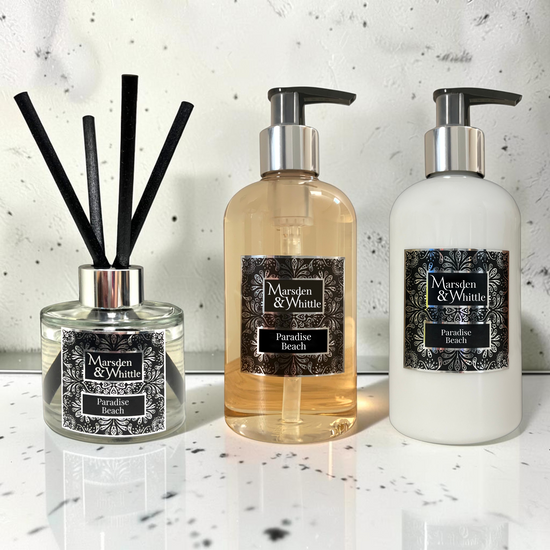 A Paradise Beach Reed Diffuser, Hand & Body Wash and Hand & Body Lotion sitting on a white marble surface with a white marble splash back behind.