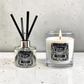 A reed diffuser and a lit scented candle from the brand Marsden & Whittle, both labeled 'Paradise Beach,' displayed on a light speckled surface against a textured light gray wall.