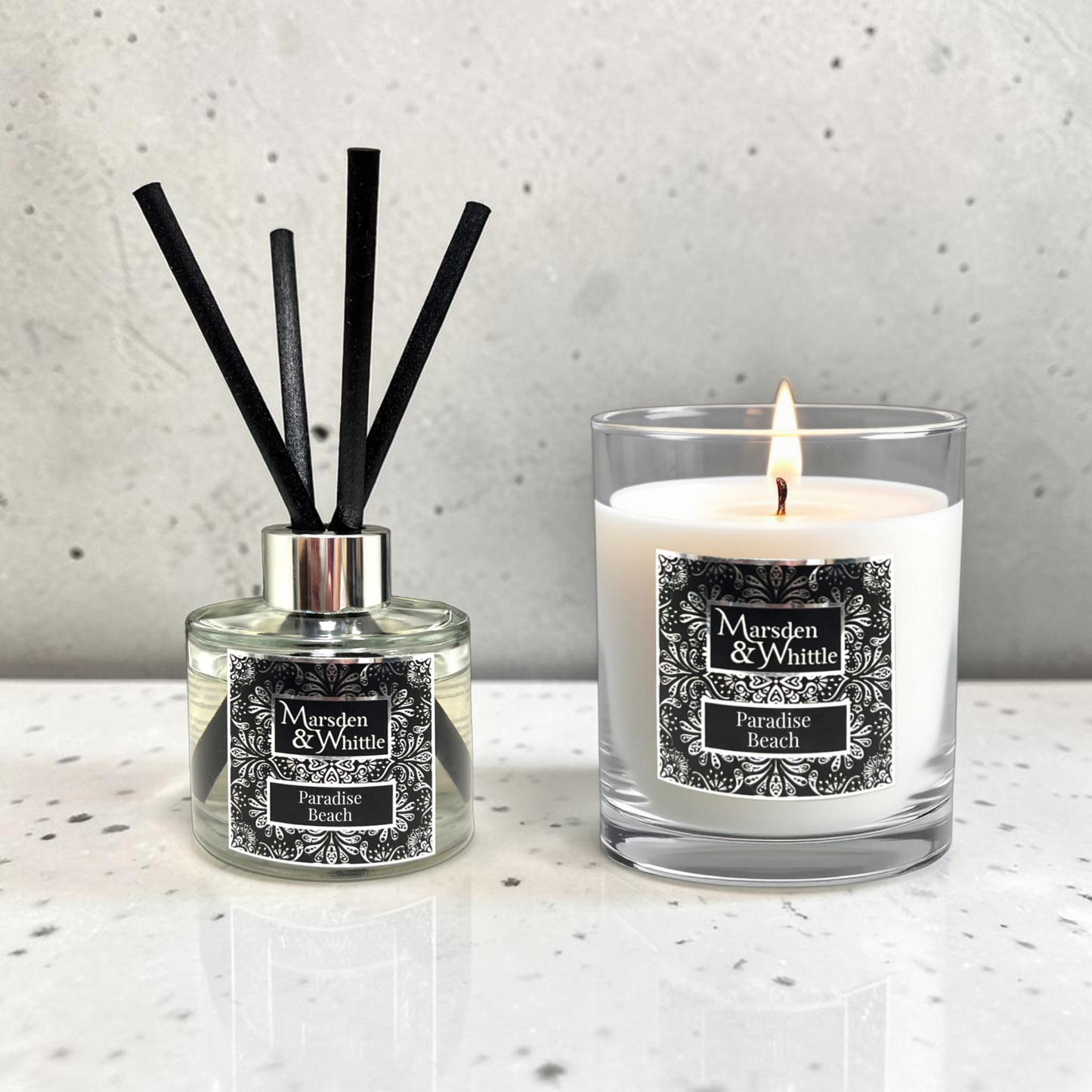 A reed diffuser and a lit scented candle from the brand Marsden & Whittle, both labeled 'Paradise Beach,' displayed on a light speckled surface against a textured light gray wall.