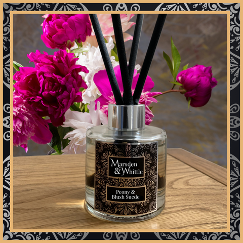 A Peony & Blush Suede glass reed diffuser with black reeds and a silver chrome cap sitting on a wooden table.
