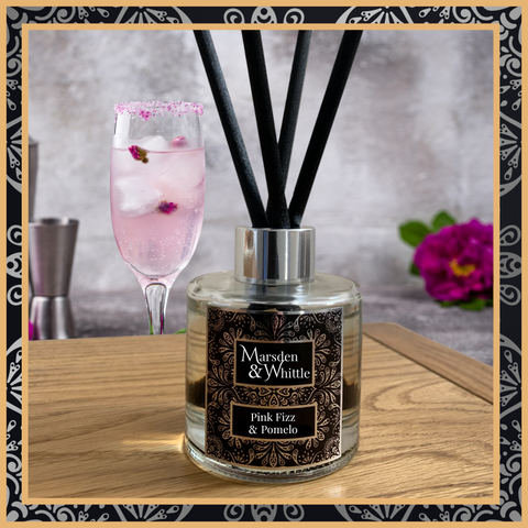 A Pink Fizz and Pomelo glass reed diffuser with black reeds and a silver chrome cap sitting on a wooden table.