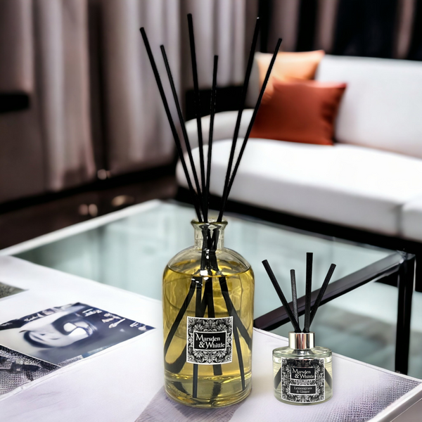 A large glass reed diffuser bottle sitting on a glass table with a cosy lounge in the background. 6 extra long black reeds are in the diffuser and the bottle is full.