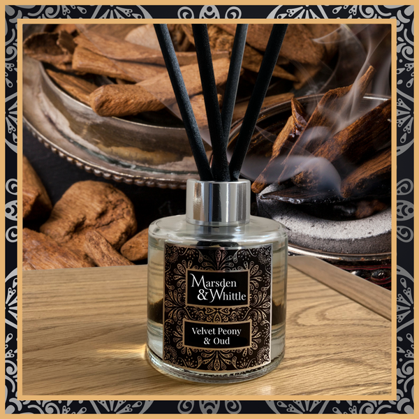 A Velvet Peony and Oud glass reed diffuser with black reeds and a silver chrome cap sitting on a wooden table.