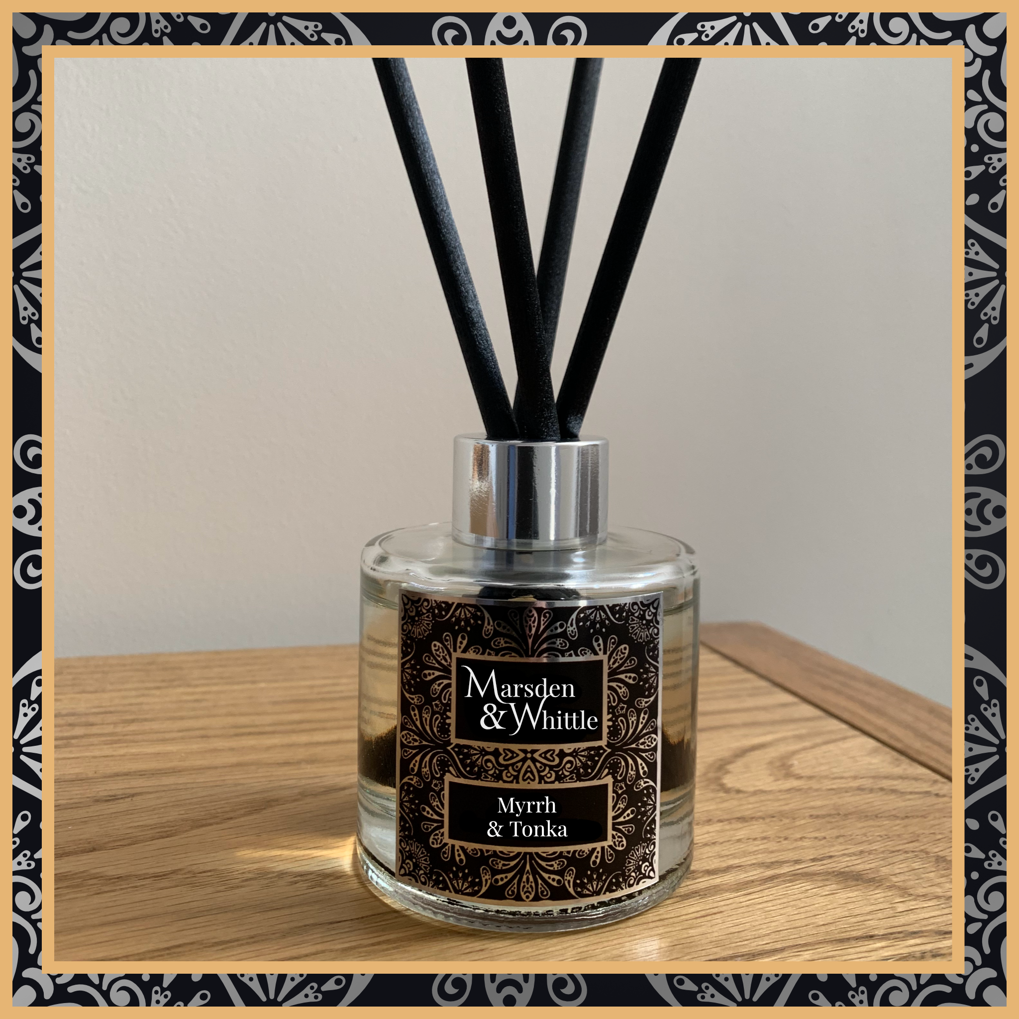 A Myrrh and Tonka glass reed diffuser with black reeds and a silver chrome cap sitting on a wooden table.