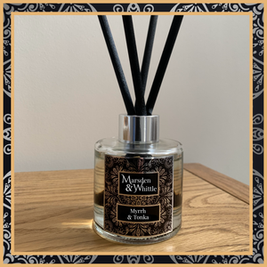 A Myrrh and Tonka glass reed diffuser with black reeds and a silver chrome cap sitting on a wooden table.