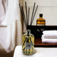 Reed diffuser with black sticks in a glass bottle on a surface, with a blurred background of towels and a bottle.