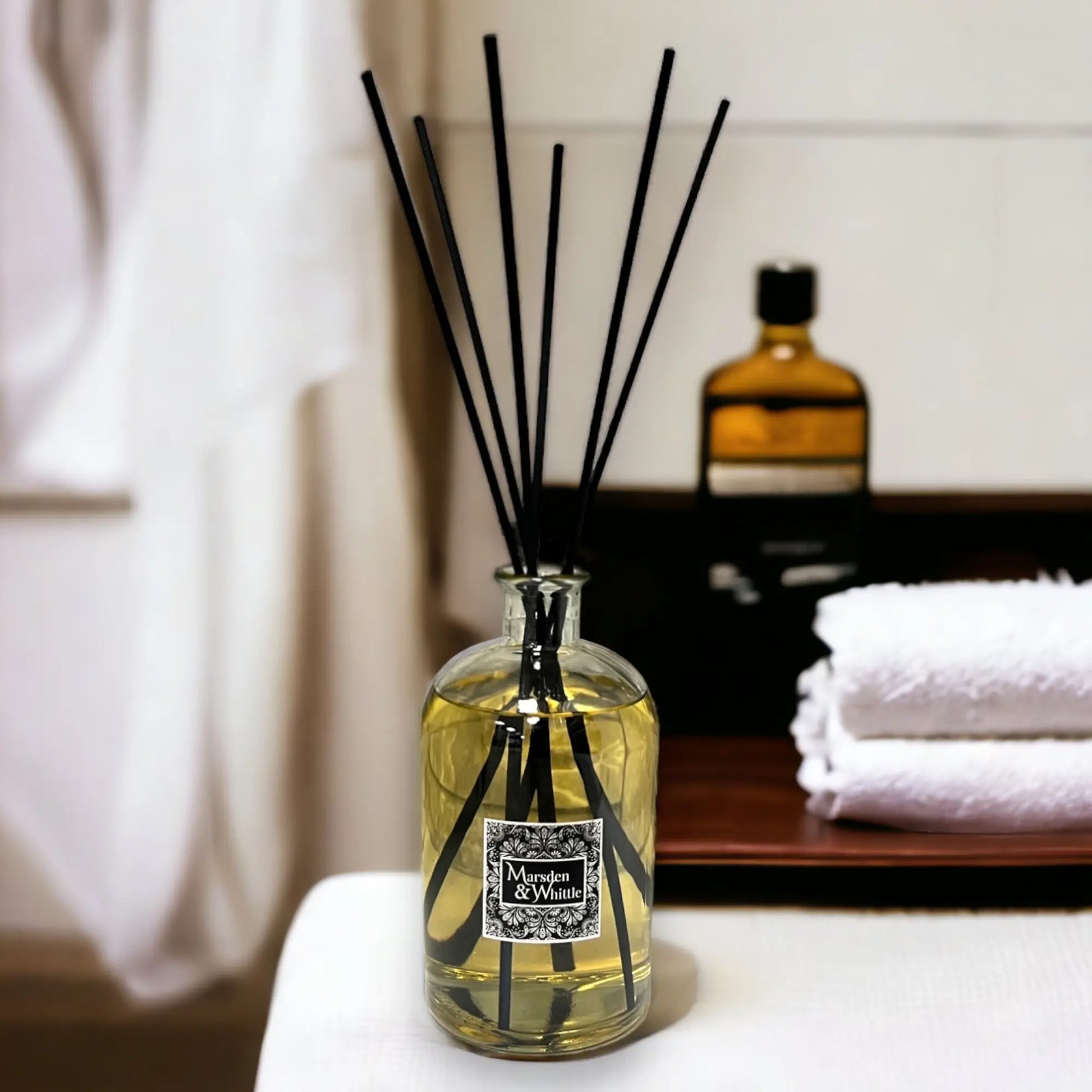 Reed diffuser with black sticks in a glass bottle on a surface, with a blurred background of towels and a bottle.