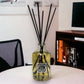 Reed diffuser in a glass bottle with black sticks on a white surface, with a blurred indoor background.