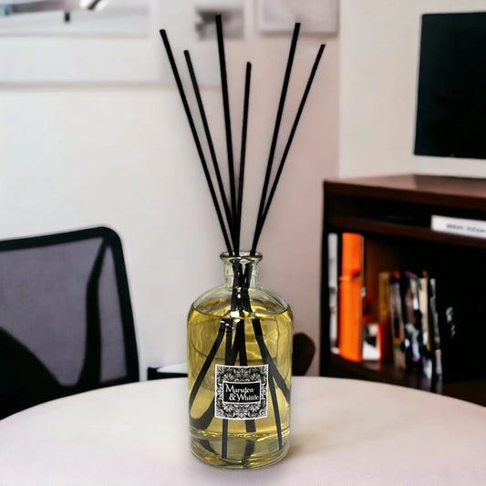 Reed diffuser in a glass bottle with black sticks on a white surface, with a blurred indoor background.