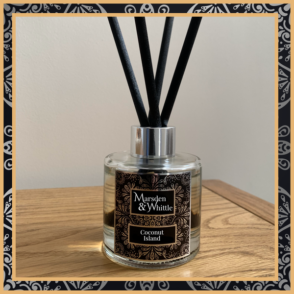 A Coconut Island glass reed diffuser with black reeds and a silver chrome cap sitting on a wooden table.