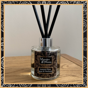 A Damson Plum, Rose and Patchouli glass reed diffuser with black reeds and a silver chrome cap sitting on a wooden table.